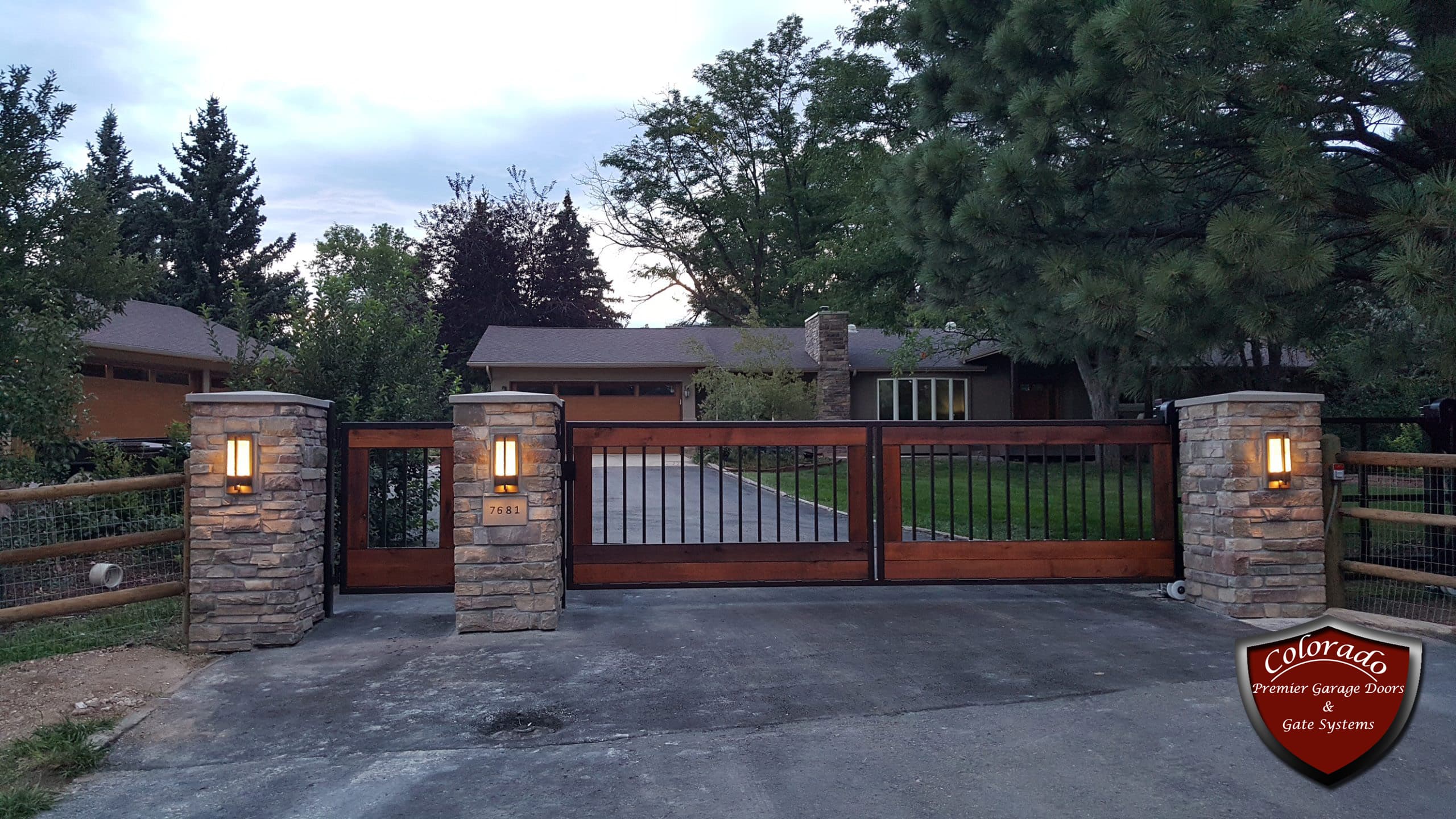 Cedar and black iron driveway gate between stone pillars with lit lamps